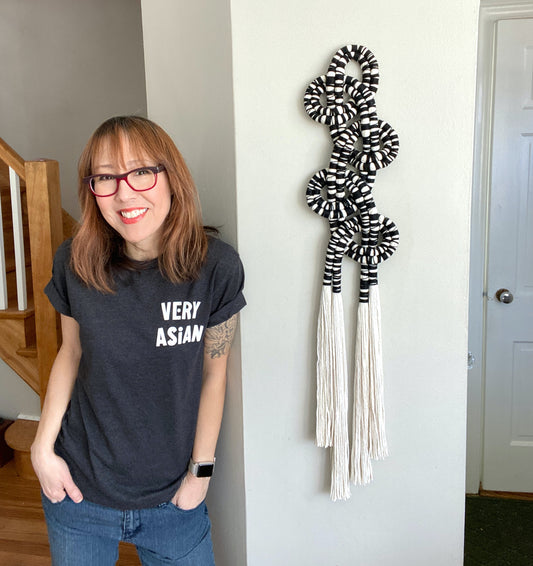 Photo of Sonya with hands in pockets & t-shirt that reads "Very Asian" standing next to a twisty fiber wall sculpture.
